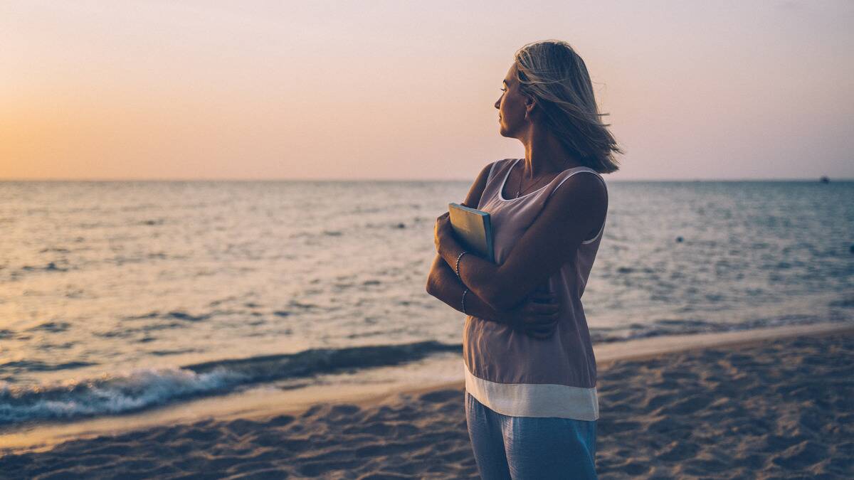 A woman standing on a beach, holding a book to her chest, looking off into the ocean. 