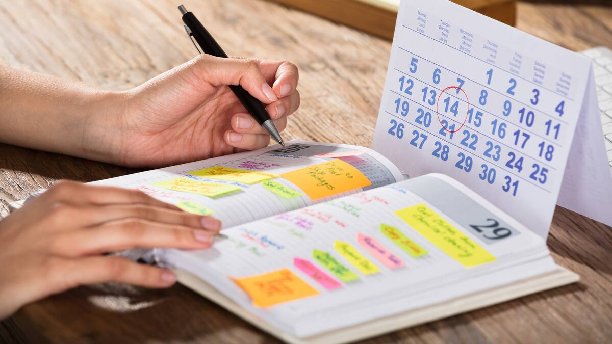 A close shot of someone filling out their schedule in a journal, a calendar on the table in front of them.