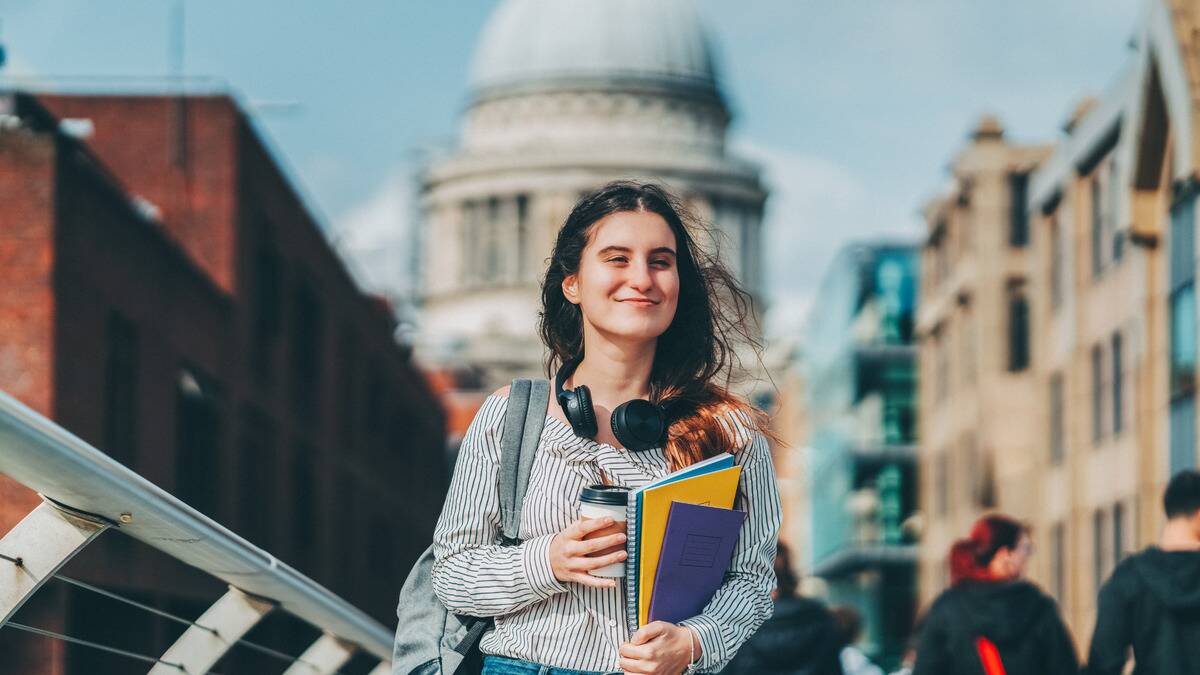 A woman smiling as she walks down the street, holding a coffee in one hand and her school notebooks in the other.