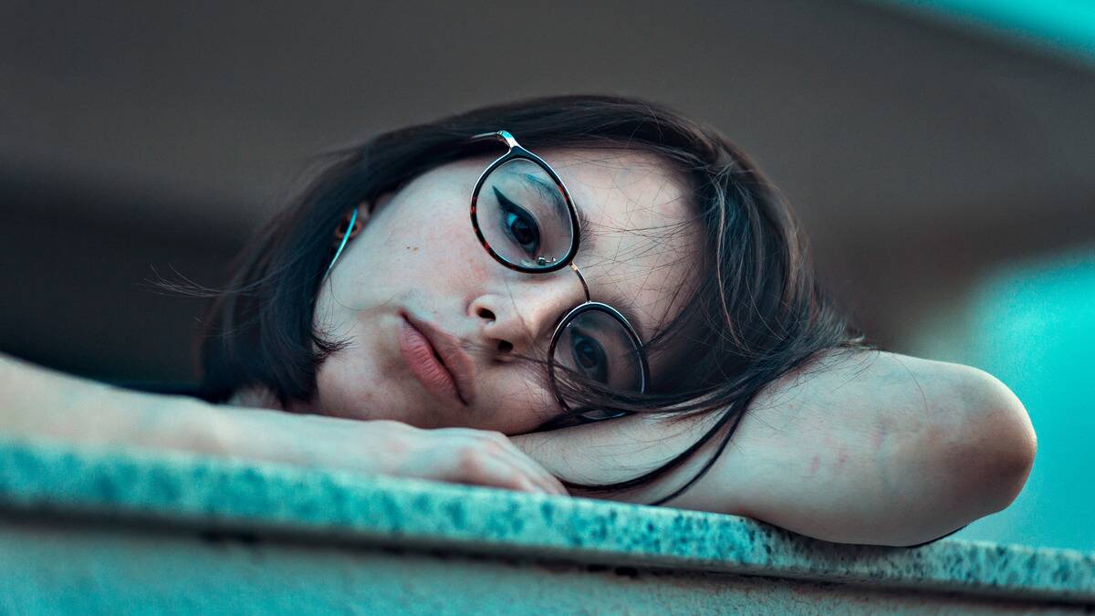 A woman resting her head upon her folded arms atop a railing, looking bored.