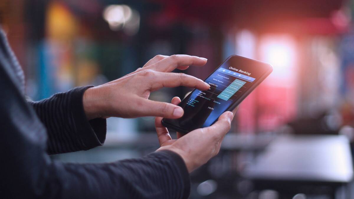 A close shot of someone holding up their phone and tapping on it, the screen showing an online banking app.