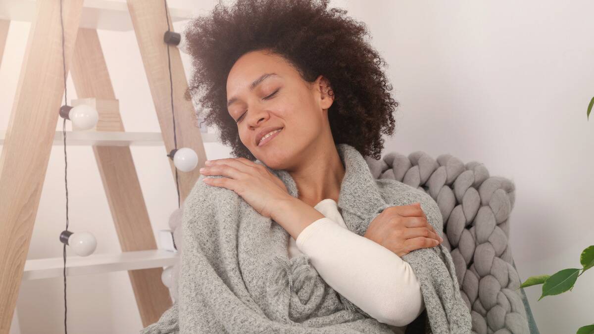 A woman sitting on a chair, wrapped in a blanket, hugging herself as she smiles with her eyes closed.