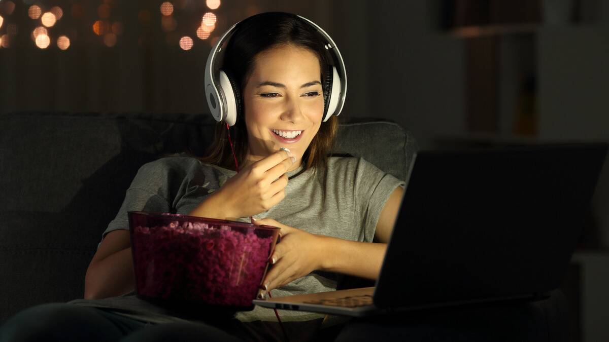 A woman sitting in front of her laptop, lit by the screen, smiling as she wears headphones and eats popcorn out of a bowl.