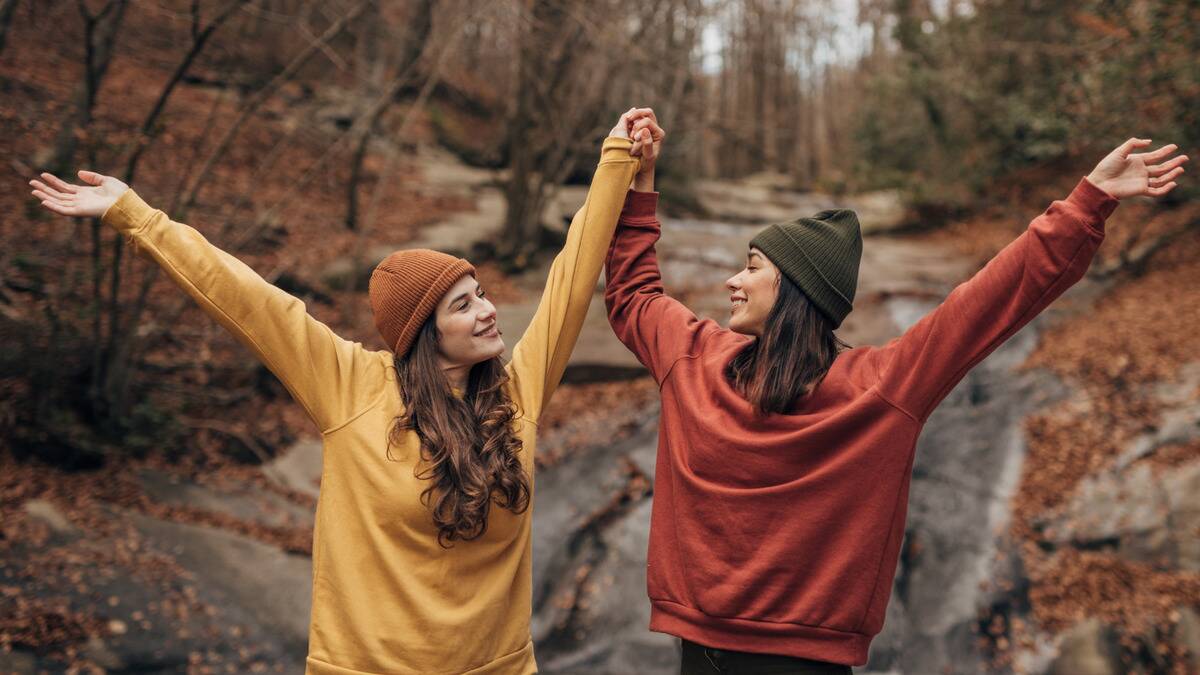 Two friends walking outside in the fall, holding hands that are raised in the air as they both smile.