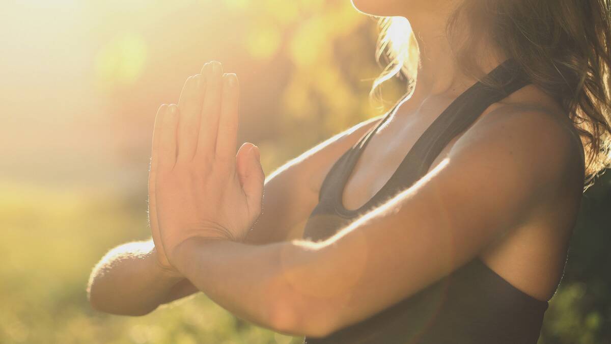 A close shot of a woman sitting outside, hands together in a prayer motion, the sun shining down on her.