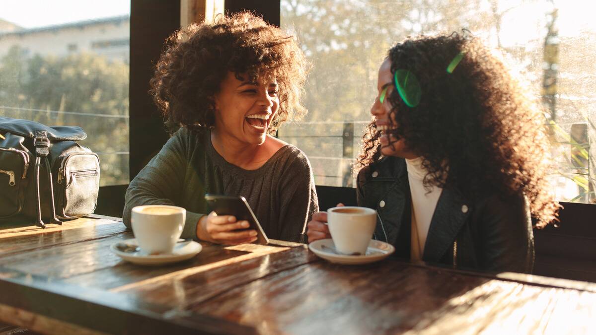 Two women sitting next to each other at a cafe, both laughing as one shows the other something on her phone.