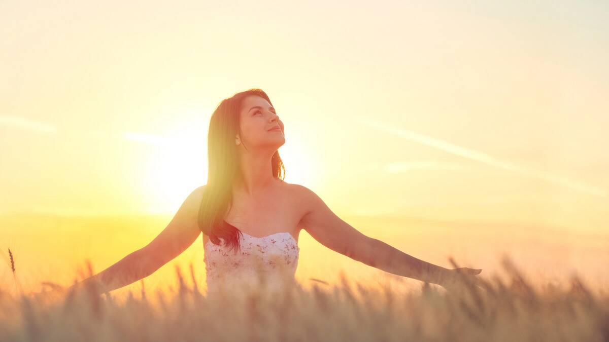 A woman standing outside in tall grass, arms out grazing the tops of it, the sun rising and shining behind her.