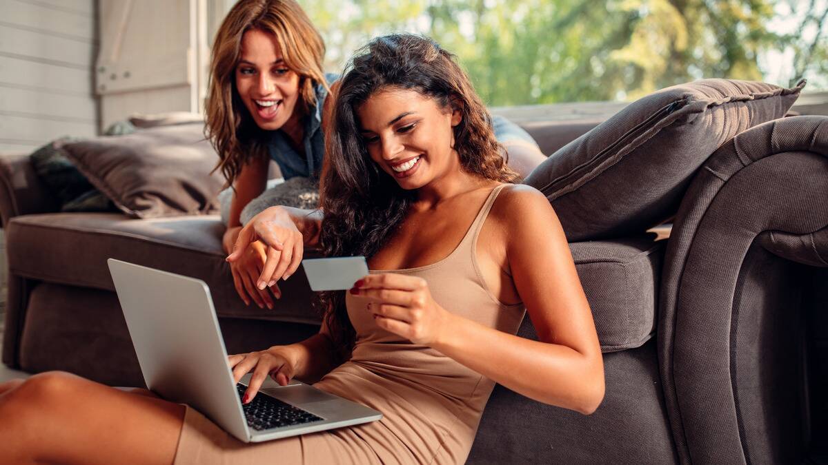 Two friends, one sitting on the floor and one laying on the couch, smiling as one buys something on her laptop with her credit card, the other friend pointing encouragingly.