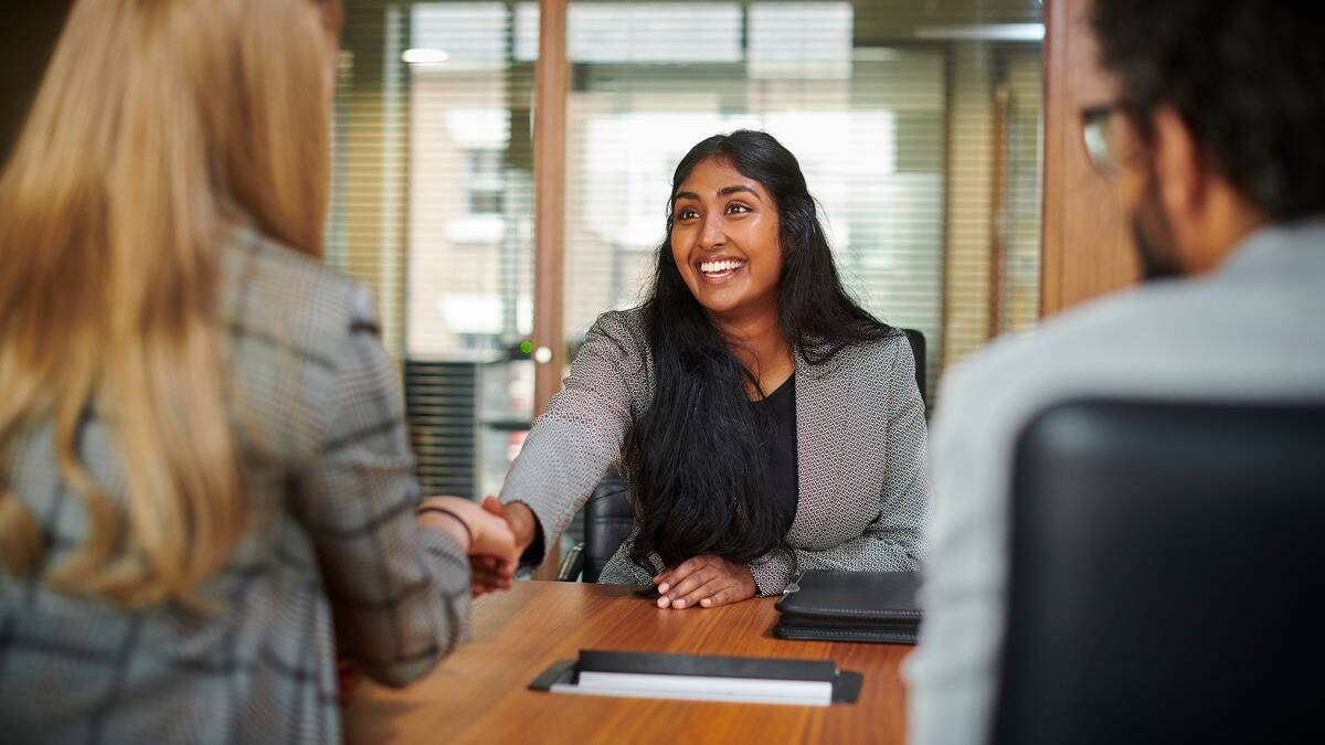 A woman smiling as she reaches across the table to shake hands with another woman, presumably for a job interview or offer. 