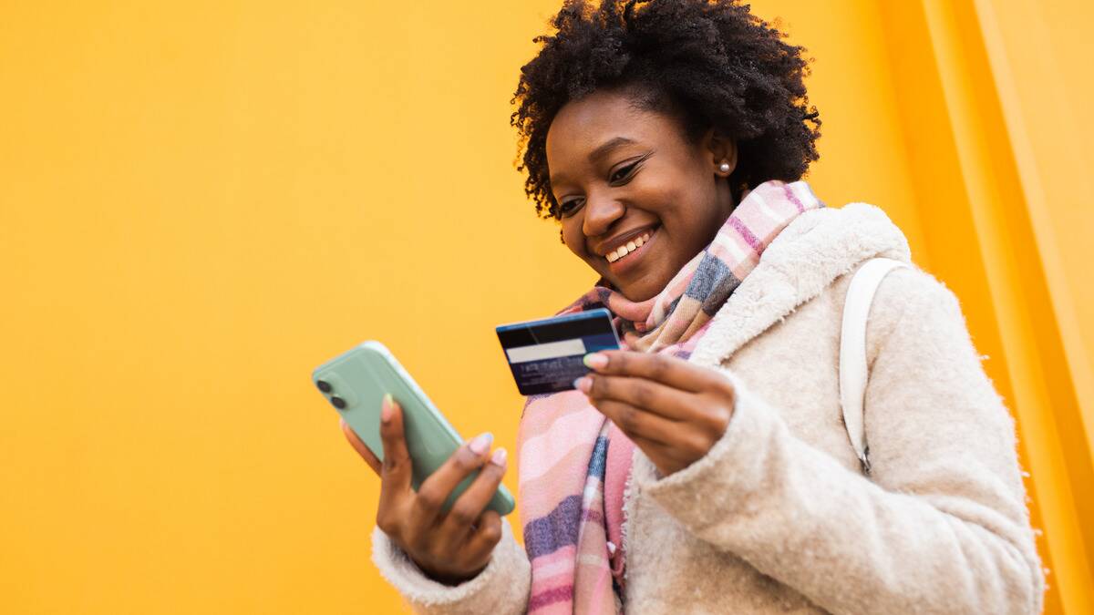 A low-angle shot of a woman looking down at her phone that's in one hand, holding up a credit card in the other, clearly purchasing something online.