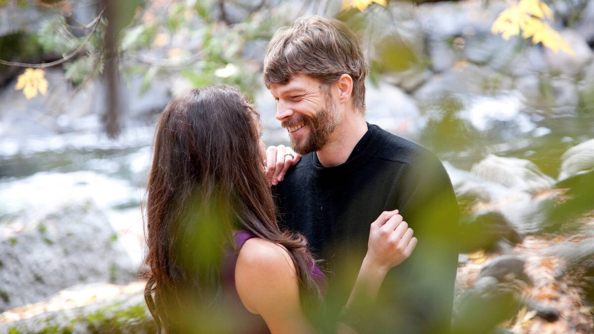 A couple embracing outside, the man smiling down at the woman, whose face we can't see, the image framed by leaves and branches that it was shot through.