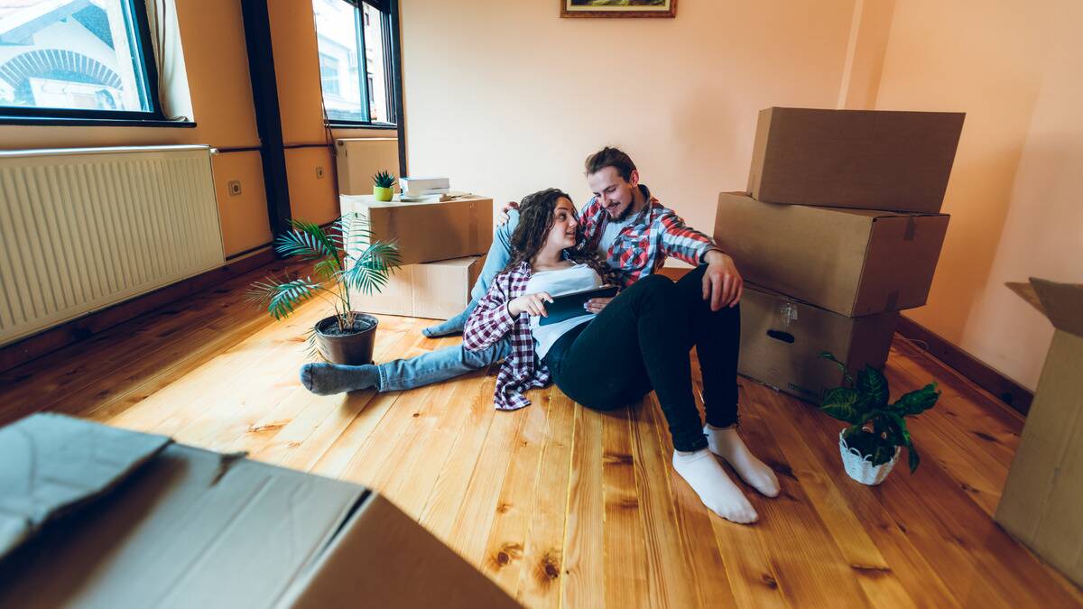 A couple sitting on the floor amid moving boxes, both looking at something on a tablet together.