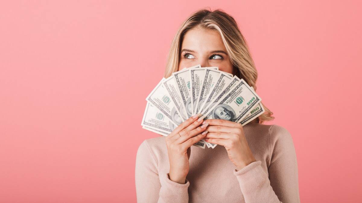 A woman against a pink background holding a fanned out stack of $100 bills in front of her face, clearly smiling as she looks off to the side.