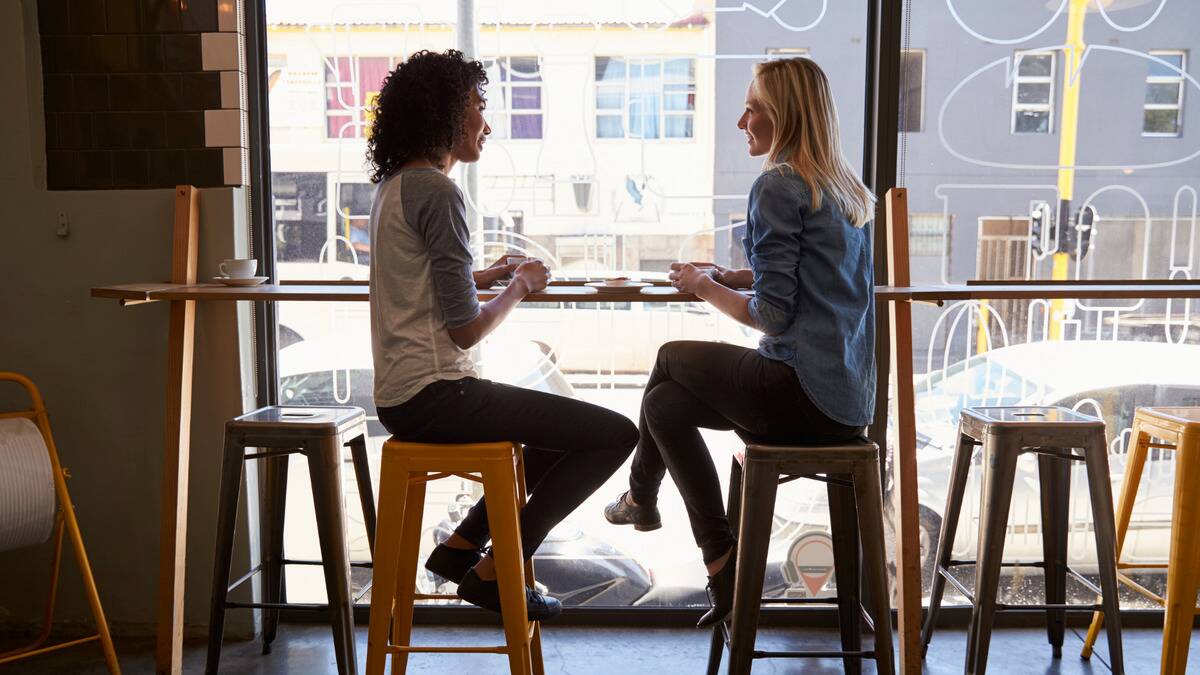 Two women sitting next to one another, turned toward each other, at a bistro, sitting at the counter in front of the window.