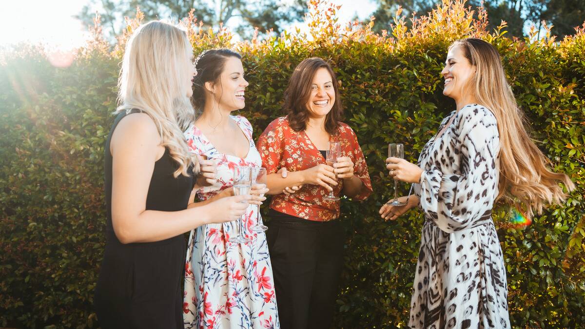 A group of friends standing outside in floral dresses, drinking champagne.