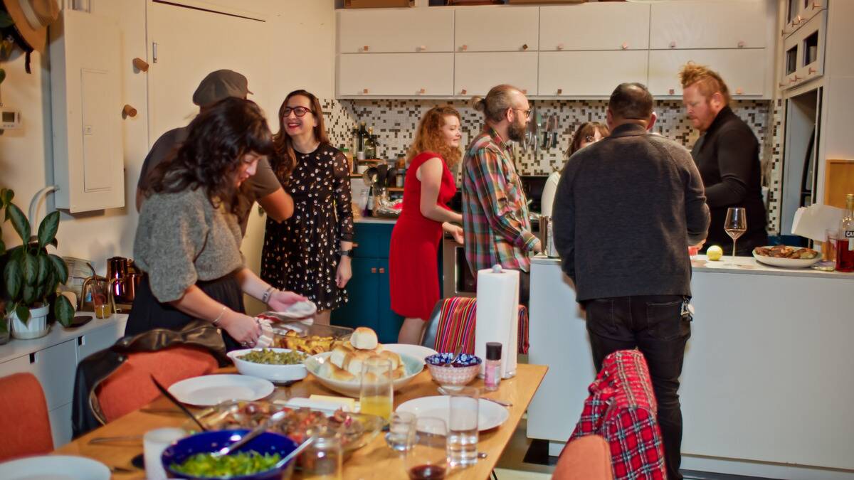 A kitchen and dining room full of guests, plates, and food during a bustling get-together.