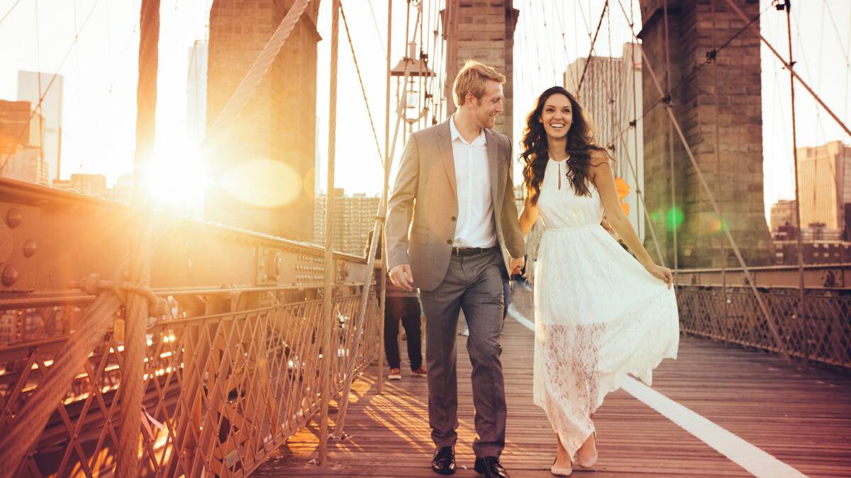 A couple talking along a bridge together, smiling brightly, the sun setting in the city behind them.