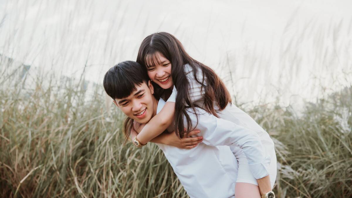 A young couple outside in a field of tall grass, both smiling as the man gives the woman a piggyback ride.