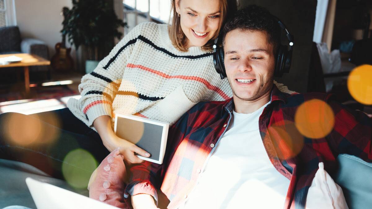 A man sitting at his laptop with his headphones on, explaining something to his girlfriend that's watching over his shoulder.