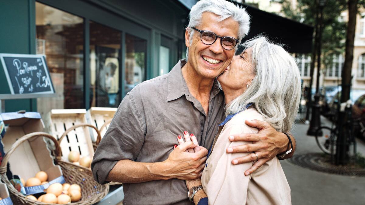 A ature couple standing outside at a market, the woman leaning up to kiss the man's cheek, putting a hand on his chest.