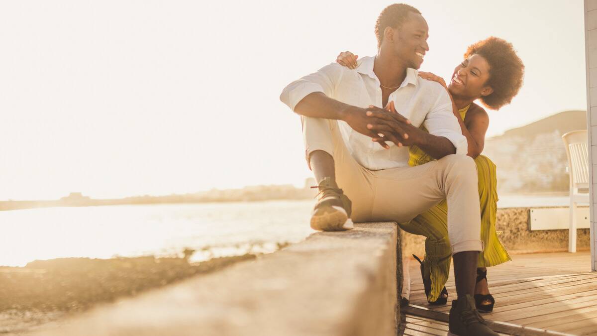 A couple sitting on a cement ledge outside, the woman behind the man with her hands on his shoulders, smiling at one another as the sun shines bright behind them.