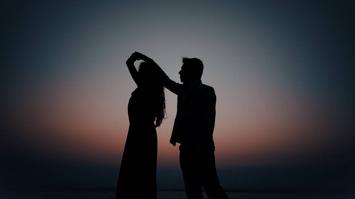 A silhouette of a couple dancing with a soft glow of the sunset behind them, the man spinning the woman under his arm.