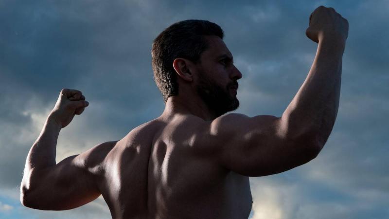 A man photographed from behind of a muscular man flexing his arms while shirtless.