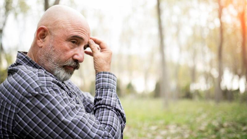 A man sitting outside, a hand to his forehead, looking thoughtful.