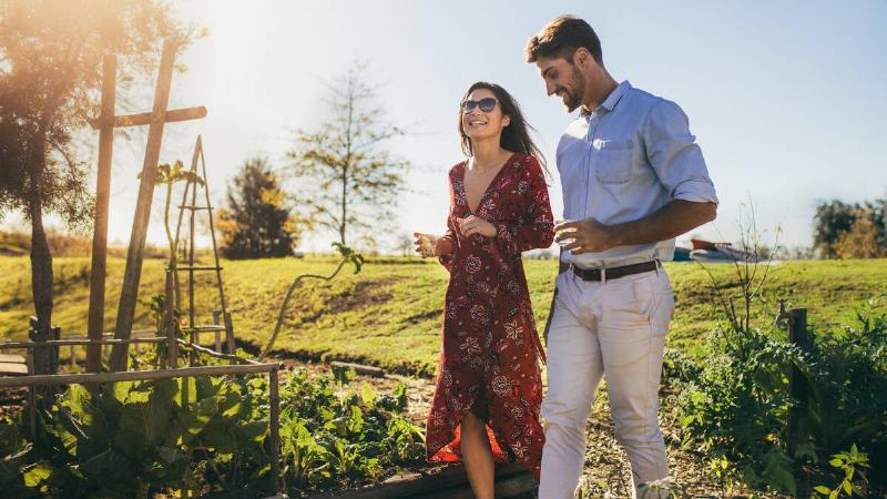 A man and a woman walking through an large garden, each holding drinks, smiling as they talk.