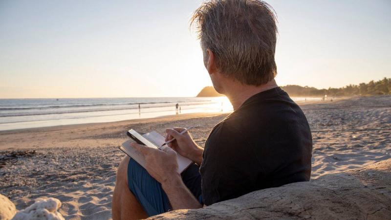 A man sitting on the beach, looking out at the horizon, writing in a journal.