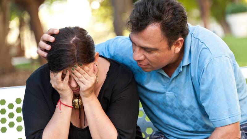 A man sitting next to his wife who's sad about something, putting an arm around her and leaning into her comfortingly.