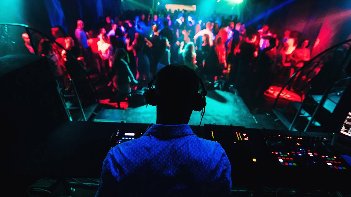 A photo from behind  a DJ at a club, a crowd dancing on the floor below him.