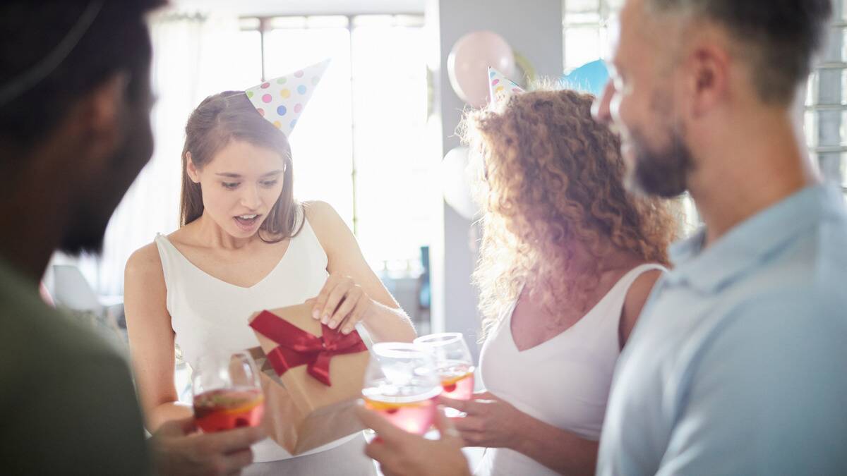 A woman looking surprised as she opens her birthday present, friends around watching her.