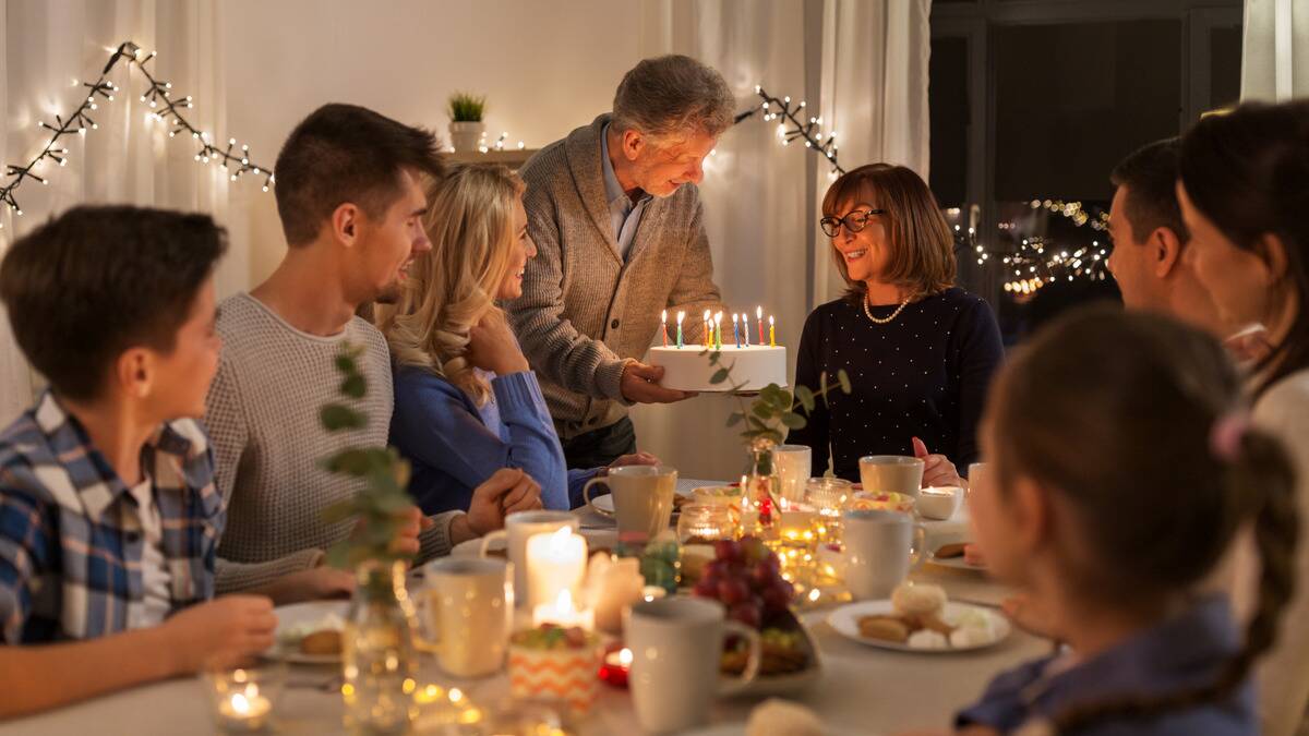 A family gathered around a table to celebrate a woman's birthday, her husband bringing her cake that's topped with lit candles.