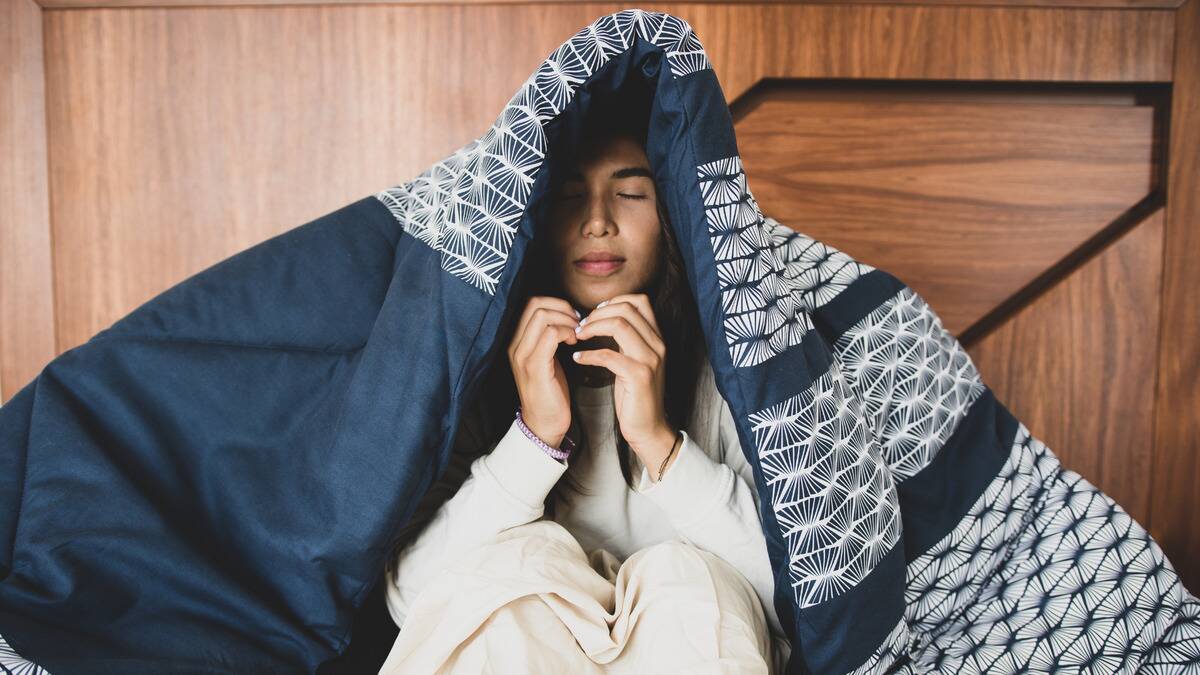 A woman sitting with her eyes closed under a comforter.