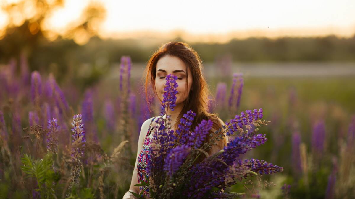 A woman sitting in a field of lilac-like purple flowers, one in front of her face.