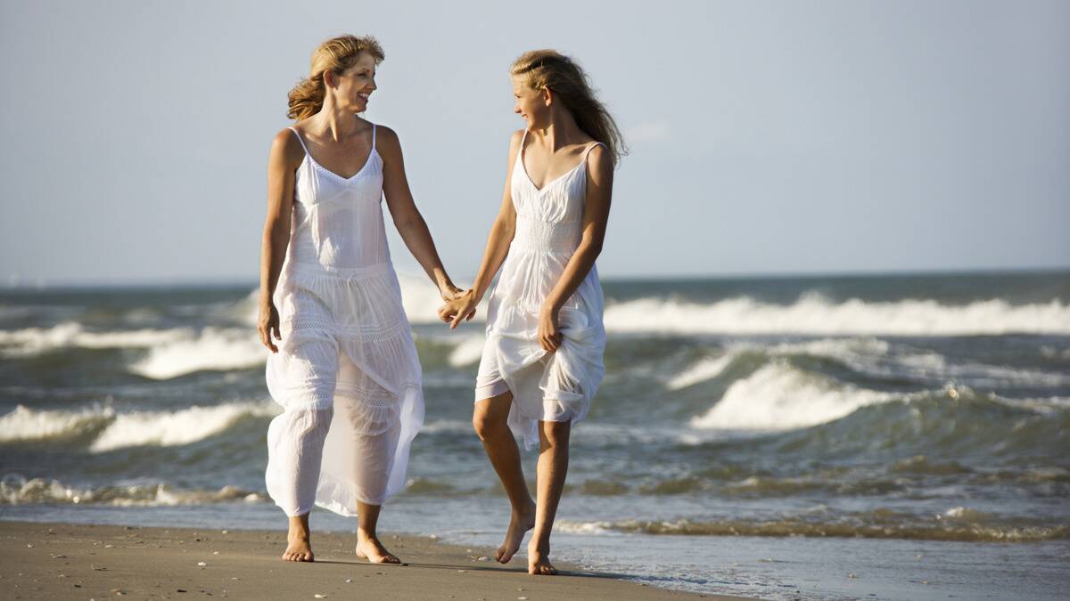 A mother and daughter, both in white flowy dresses, holding hands and smiling at each other as they walk along the beach.