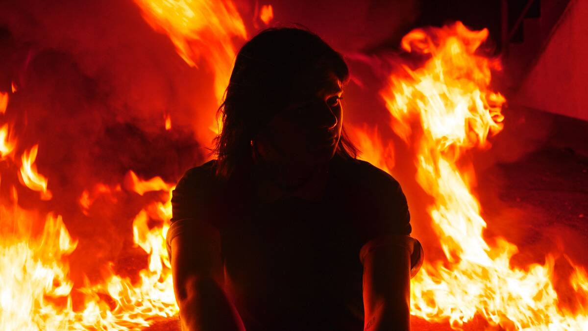 A woman kneeling outside, fire raging behind her.