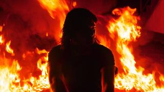 A woman kneeling outside, fire raging behind her.