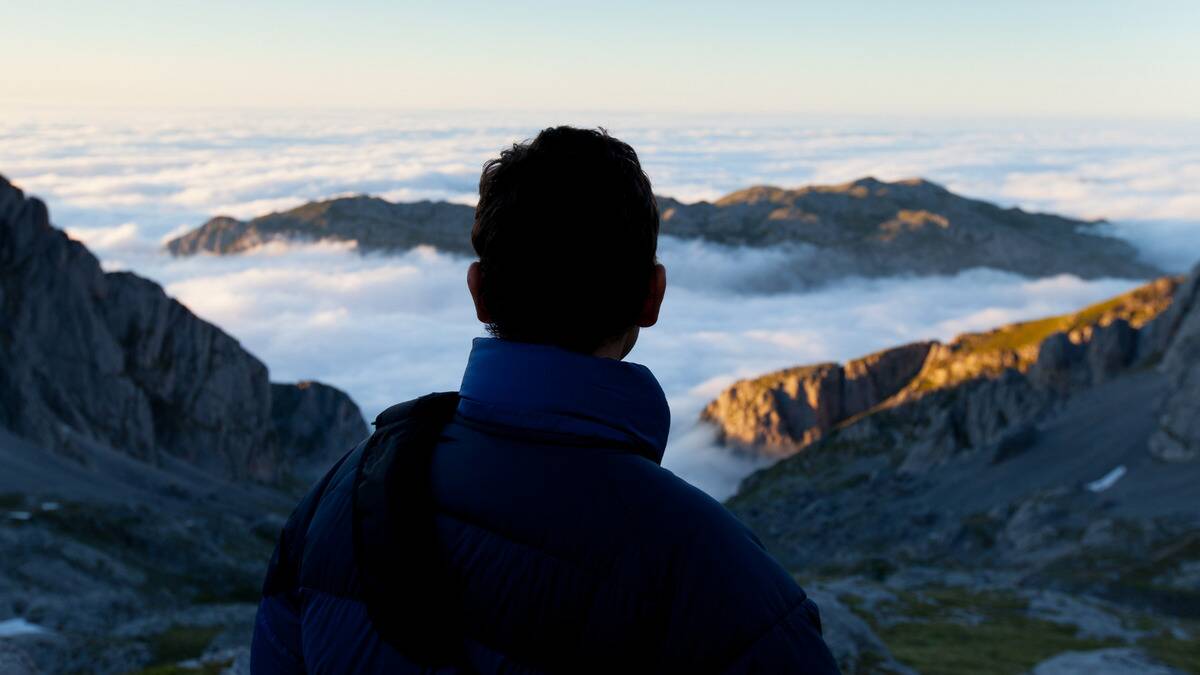 A photo from behind of a man upon a mountain, looking over at the range of clouds with other mountain tops cresting through.