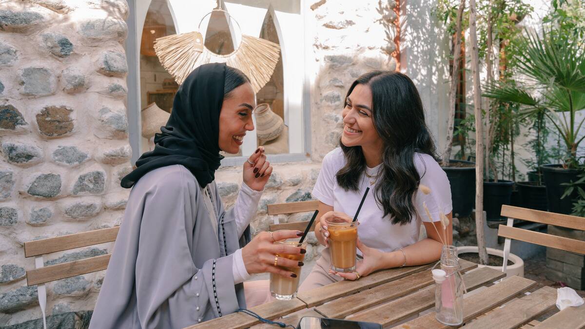 Two friends sitting side by side at a cafe but turned toward each other, smiling as they chat.