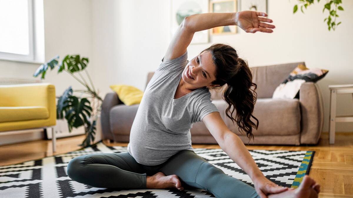 A woman sitting on the floor of her living room, one leg folded in, the other outstretched, stretching as she reaching both arms toward the further leg, one reaching over her head.