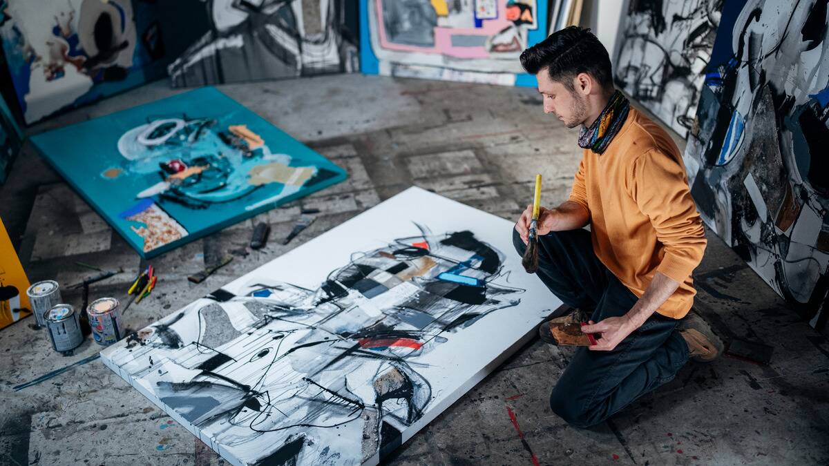 A man kneeling on the ground next to a large canvas that he's painting on.