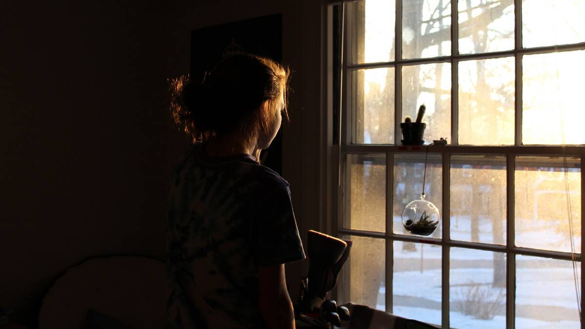 A girl standing in a dark room looking out a window, the sun shining on her face, showing us the outline of it.