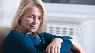 A woman sitting on a couch, a knee drawn up, both of her arms folded onto it, looking down with a sad and weary expression.