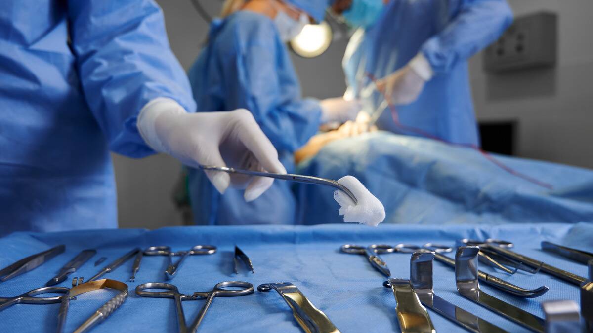 A low shot of a table of surgical tools, one person reaching to place a piece of gauze on the table. There's an active surgery happening blurred in the background.