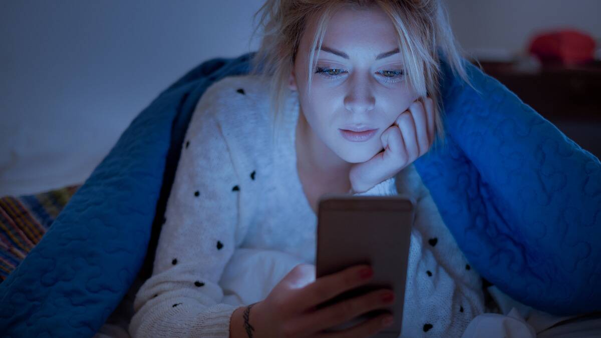 A woman laying on her stomach in bed, holding her phone in one hand, her chin in her hand.