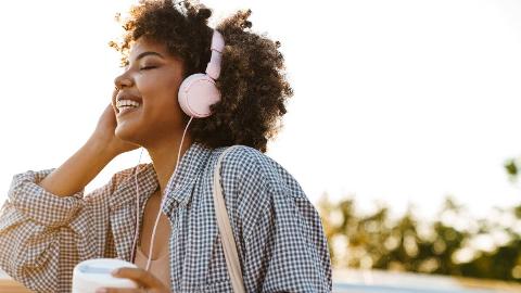 A woman walking outside with headphones on, smiling with her eyes closed as she listens to her music, one hand raised to touch the cup of her headphones.