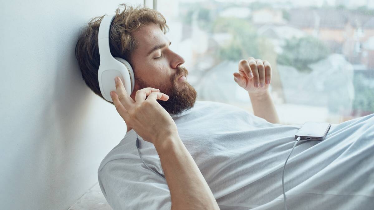 A man lounging by a window with headphones on, eyes closed, one hand raised to touch the cup of his headphones.