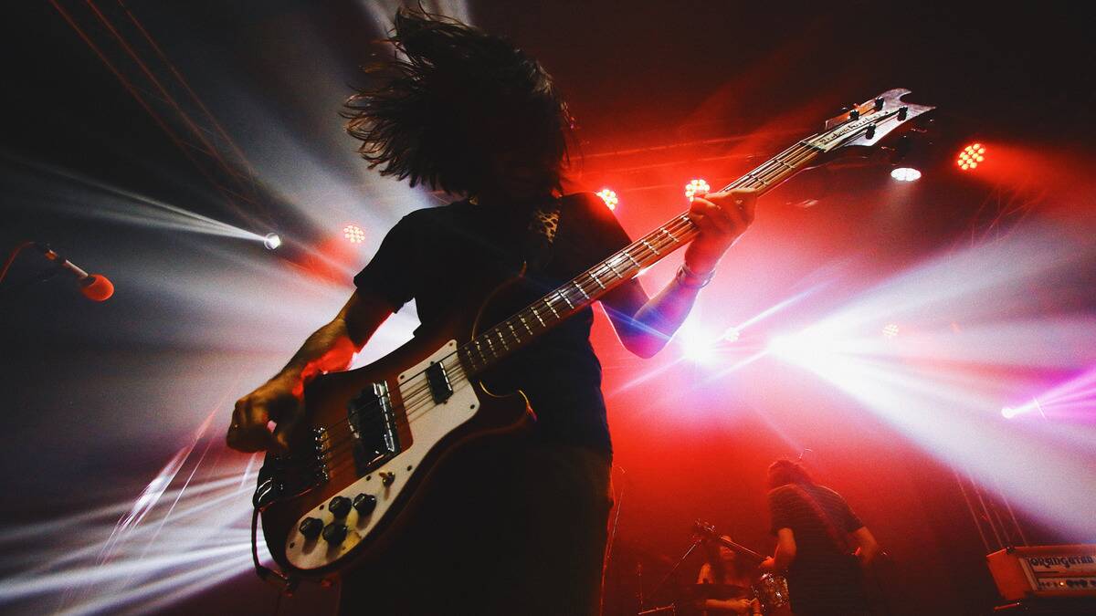 A close shot of a man playing a bass guitar on stage during a show, bent over so his hair is in front of his face.